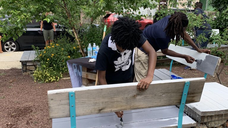 two teenager paint and tend to benches in a park surrounded by a garden