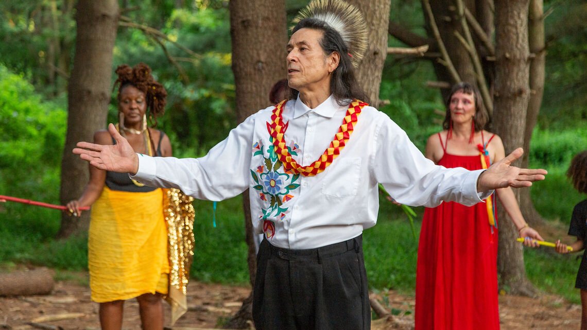 Three figures in the frame, one man in the center flagged by two women behind him. The man's arms are outstretched.