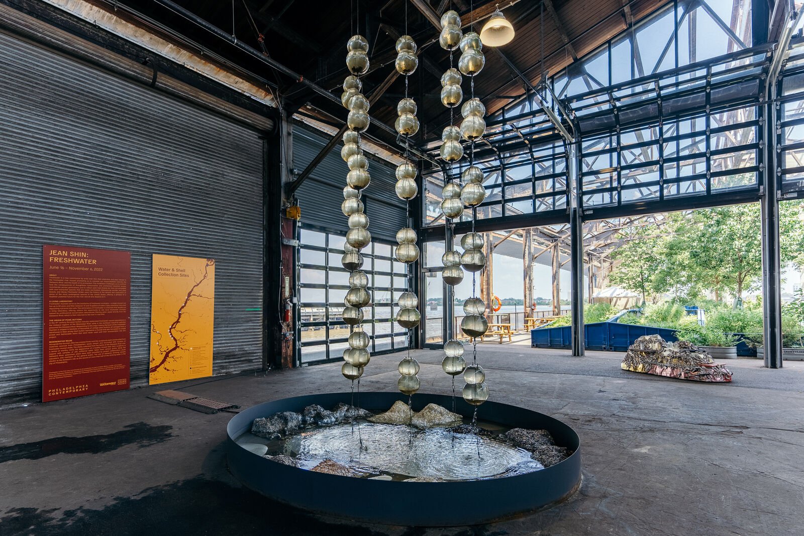 In an open air pier, bulbous glass vessels are suspended in the air from top to bottom. There are four rows of glass vessels, a few contain live mussels that are filtering water in real time. Below the suspended vessels is a shallow fountain basin filled with water trickling down from above, which has been filtered by the mussels. In the basin are pearl blankets in various mounds, the pearls range from snowy white pearlescent to the darker outer shell color of mussels. The pearl blankets were hand sewn and shimmer with each drop of water that comes into contact with them.