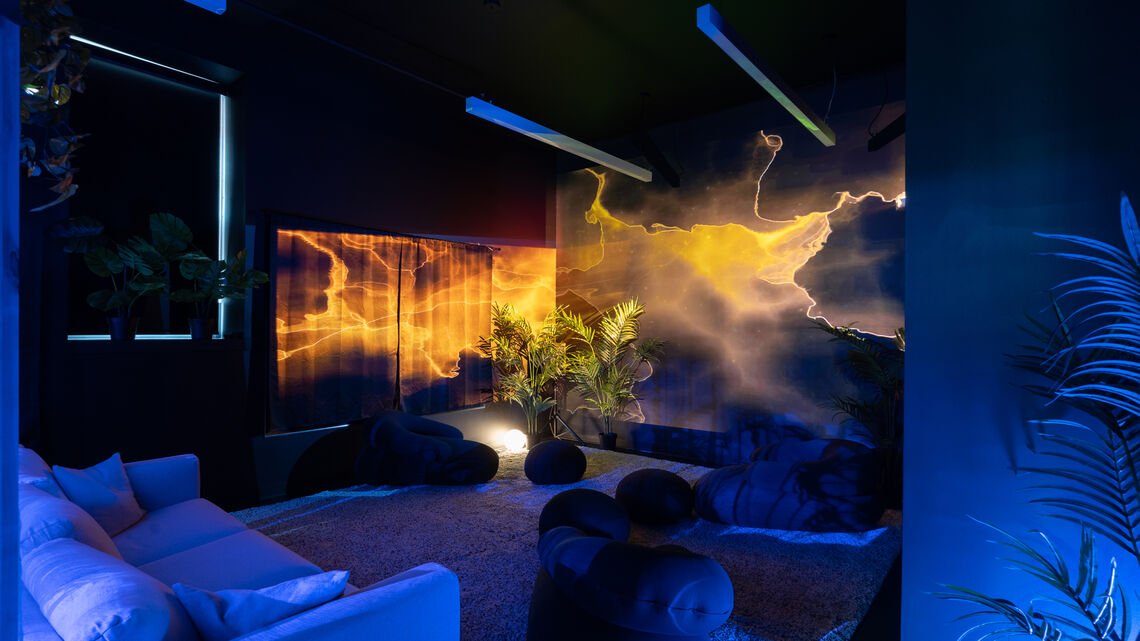 Interior of Supine Horizons exhibition space. Light and visuals are projects onto the walls on the right-hand side, colors are orange and blue. A large white couch is on the left-hand side, smaller dark round cushions are placed in the middle of the room. The leaves of a palm plant peek out on the right corner of the frame.